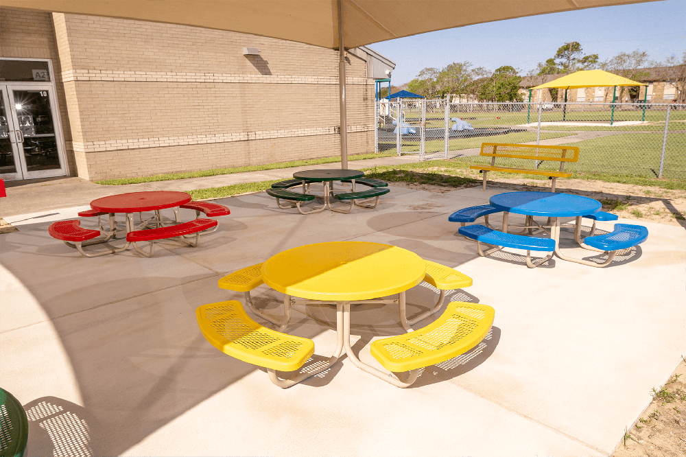 Colorful round children’s picnic tables in red, yellow, blue, and green installed under a shade canopy at an elementary school playground.